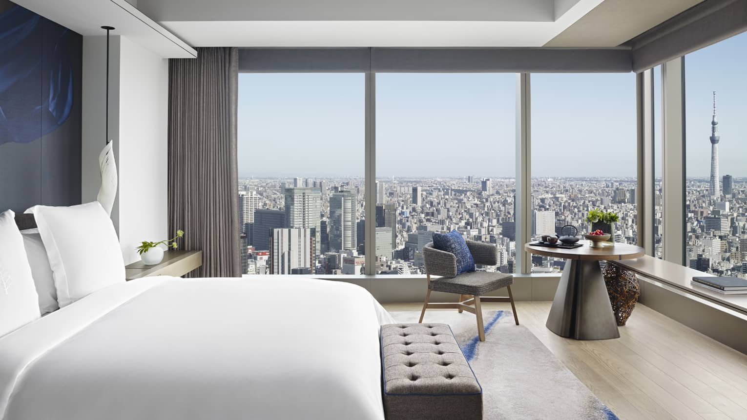 Guest room with king bed, tufted bench, round table with armchair and floor-to-ceiling windows offering city views