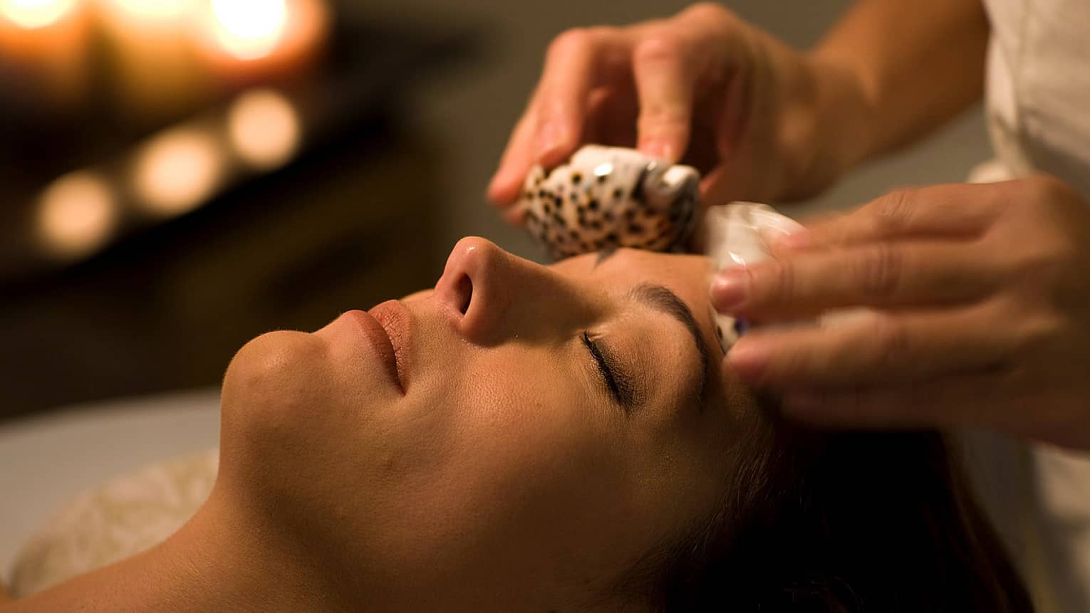Close-up of woman on table at Seaside Spa with eyes closed, attendant placing sea shells above eyebrows