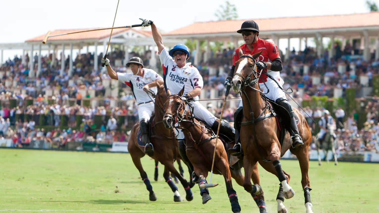 Three men with polo mallets ride horseback across field, large crowds in stand behind them