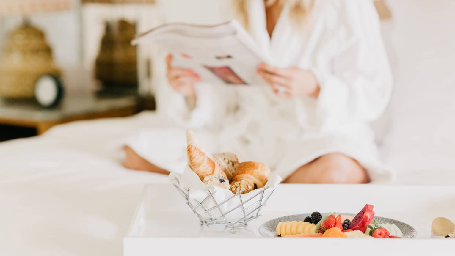 Pastries, fruit on in room dining tray, woman wearing white bath robe reads in background