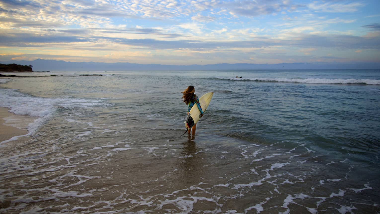 Woman with white surfboard walks into ocean tide at sunset