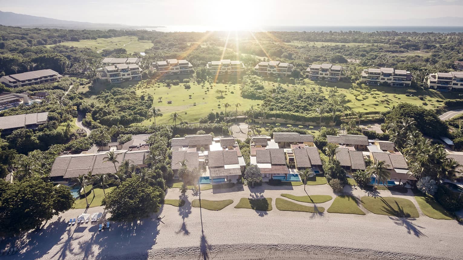 Aerial view of green lawns, trees and estate rooftops behind white sand beach at sunset