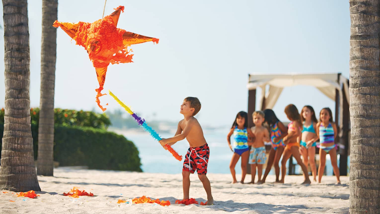 Boy in bathing suit on beach swings stick at orange pinata, children watch in background
