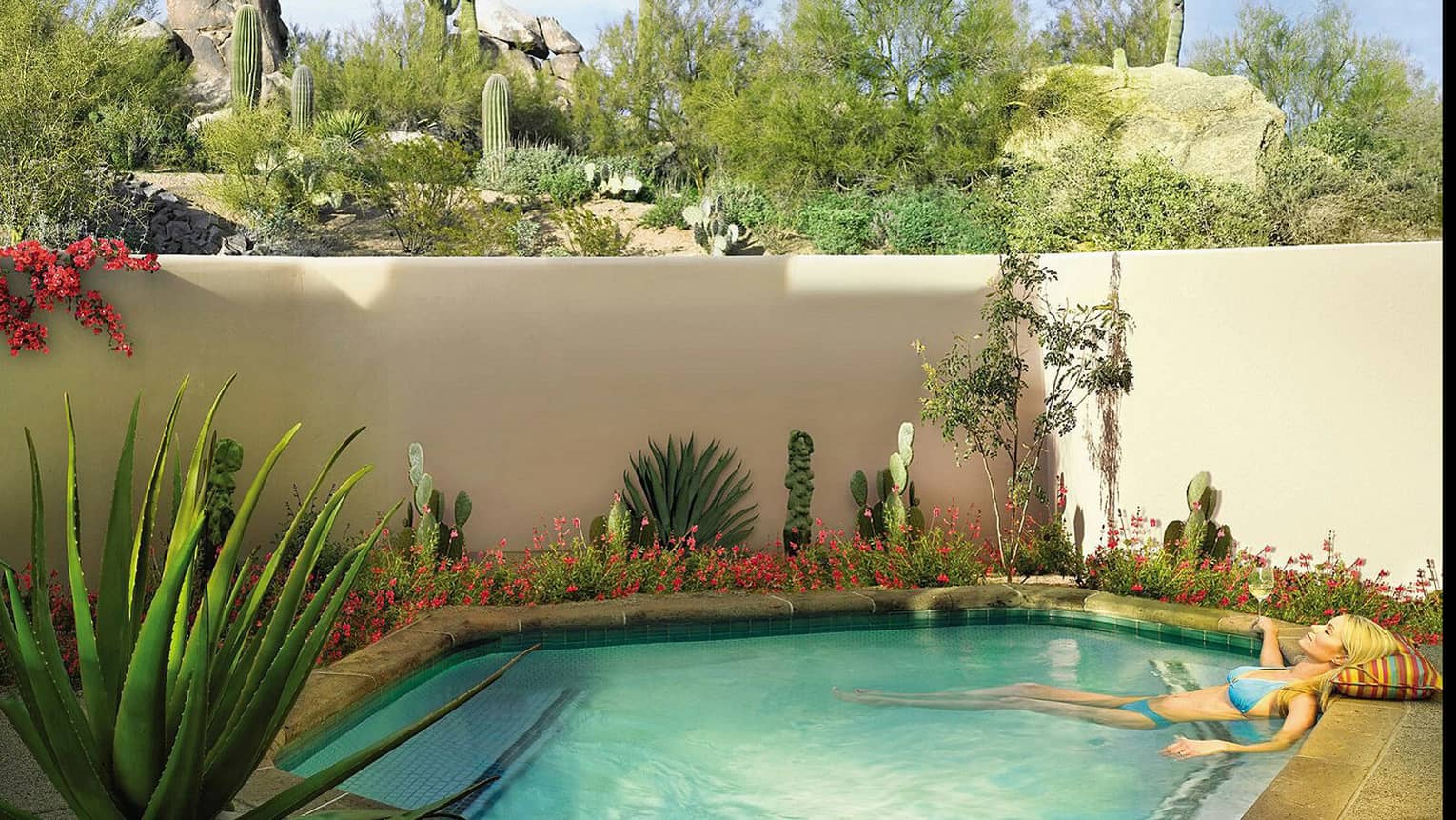 Woman wearing blue bikini wades in plunge pool with glass of white wine, cactus plants under white wall
