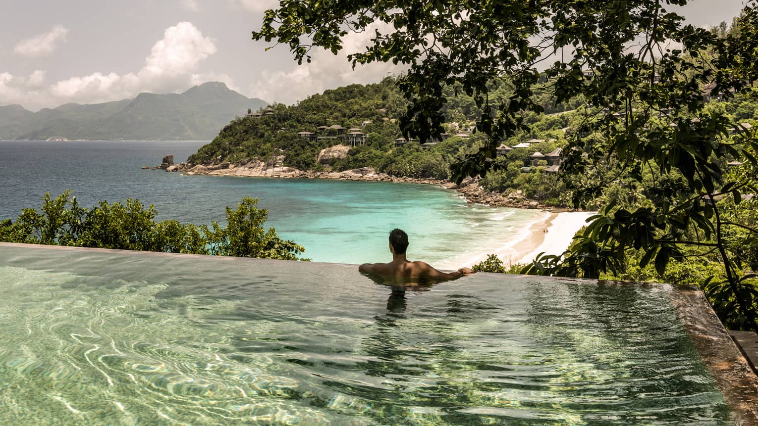 Back of man shoulder-deep in infinity pool, looking below at resort beach, ocean