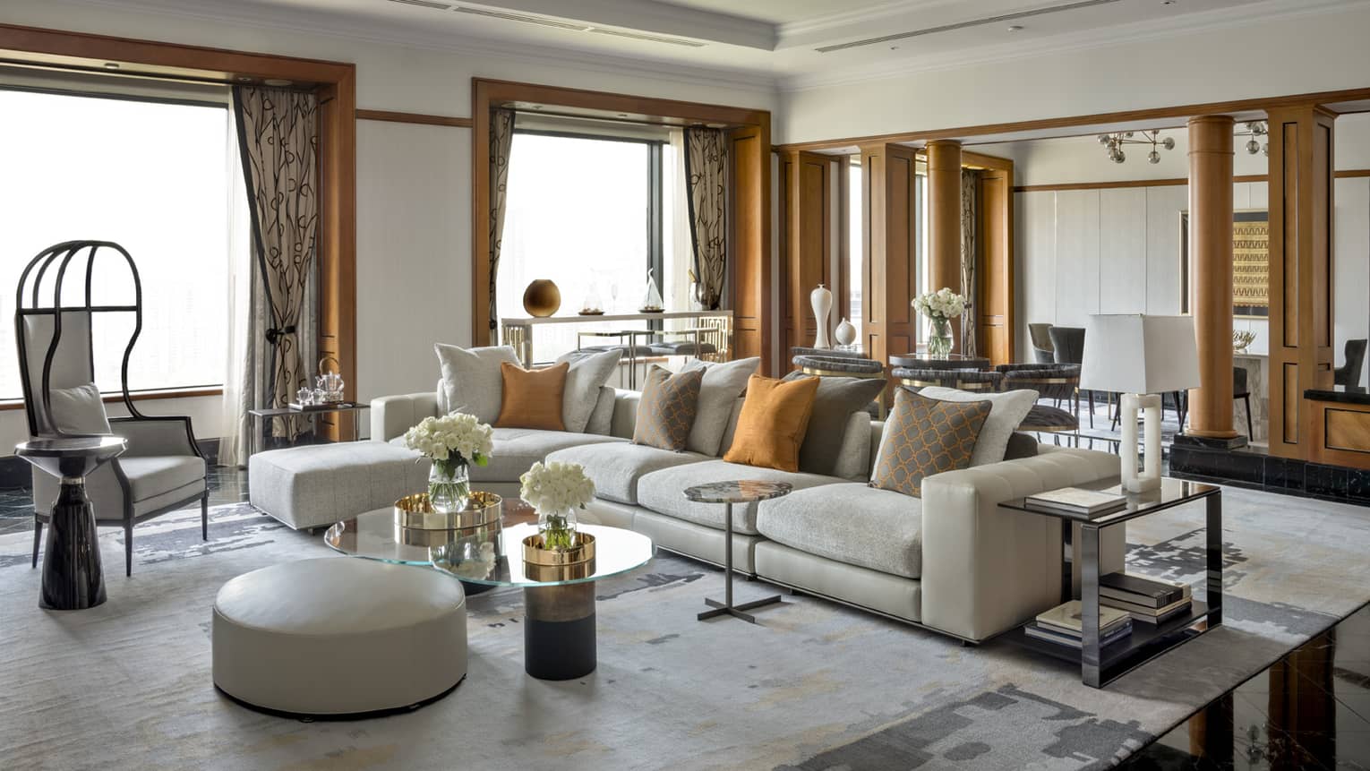 A spacious, luxurious living room with large sectional sofa, orange accent pillows, modern chairs and glass-topped tables. The room features wooden accents and natural light.
