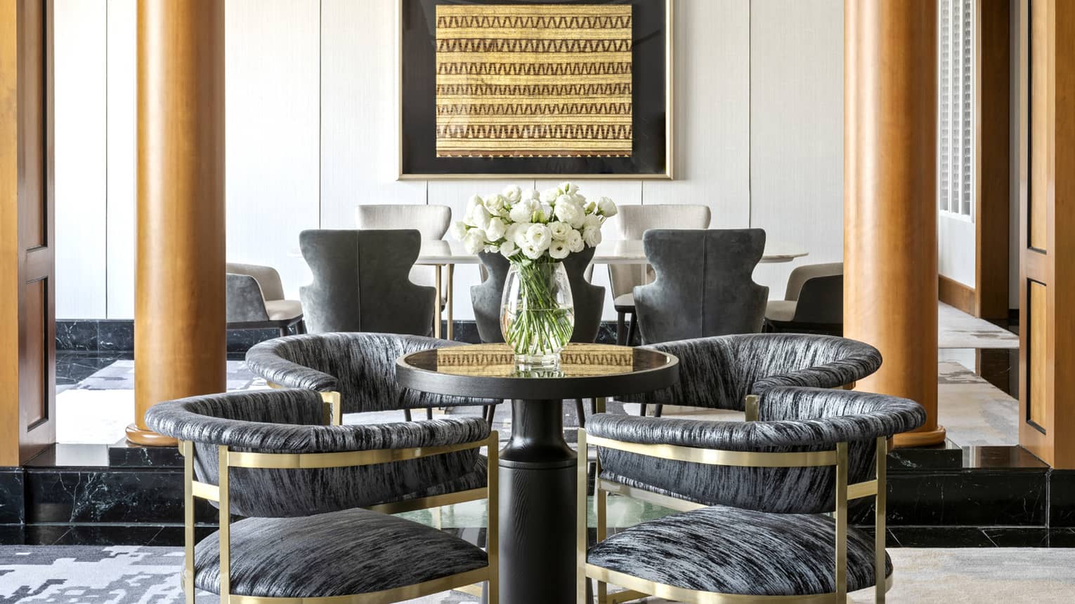 A stylish dining area featuring a small round table with modern cushioned chairs and a vase of white flowers. The space is accented by wooden columns and framed wall art.