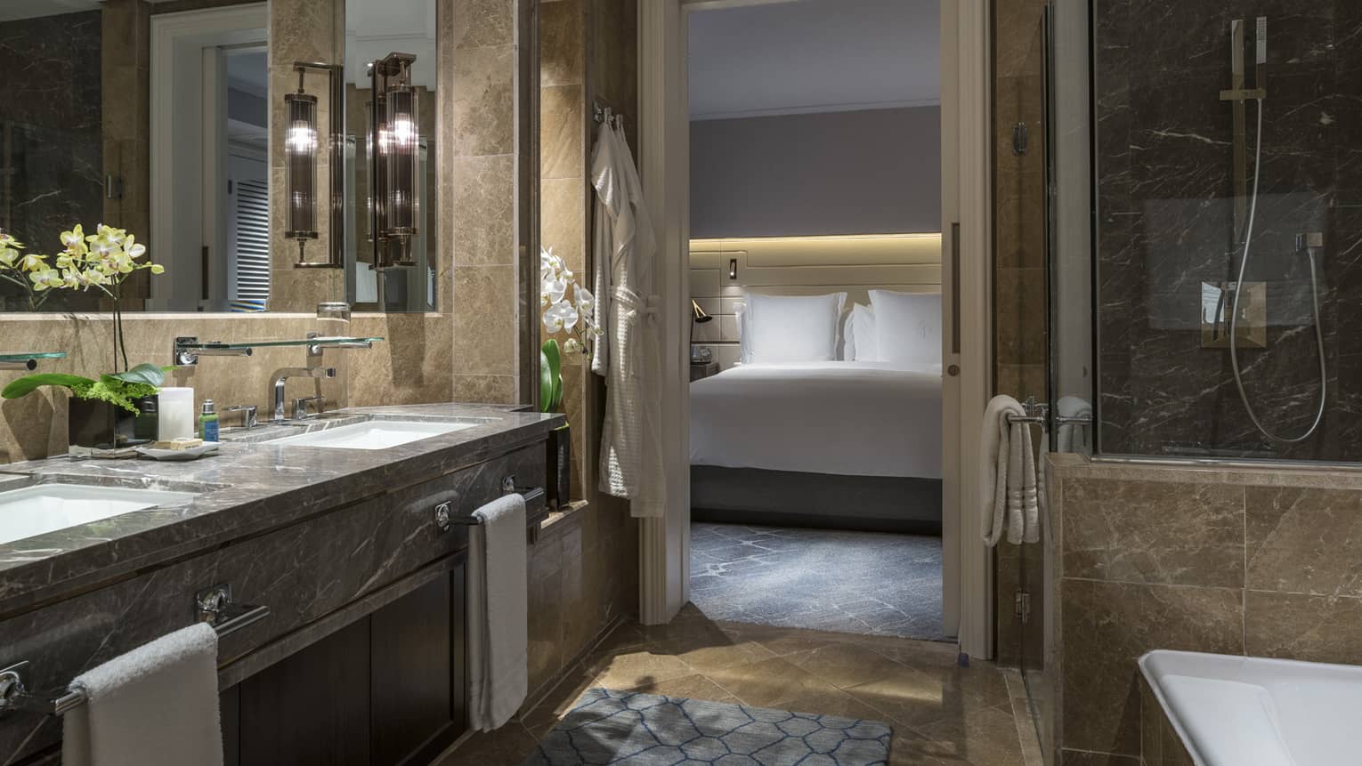 A marble bathroom with double sinks, glass-enclosed shower and bathtub. The bedroom, featuring a well-lit bed with white linens, is visible through a sliding door.
