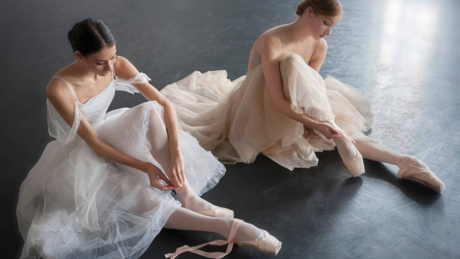 Two ballerinas in long dresses sit on floor, lace up ballet shoes at Mikhailovsky Theatre