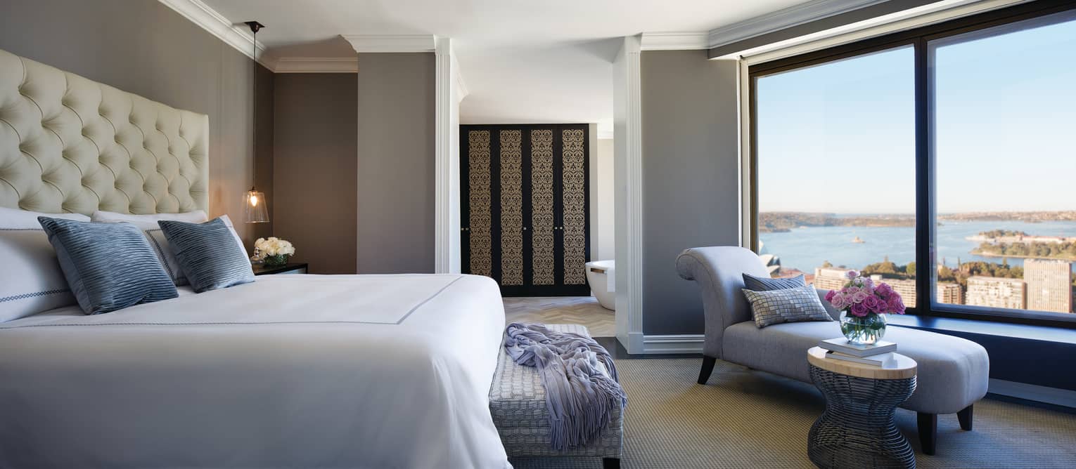 Royal Suite bed, scarf over footstool, blue chaise, table, flowers by window with ocean views