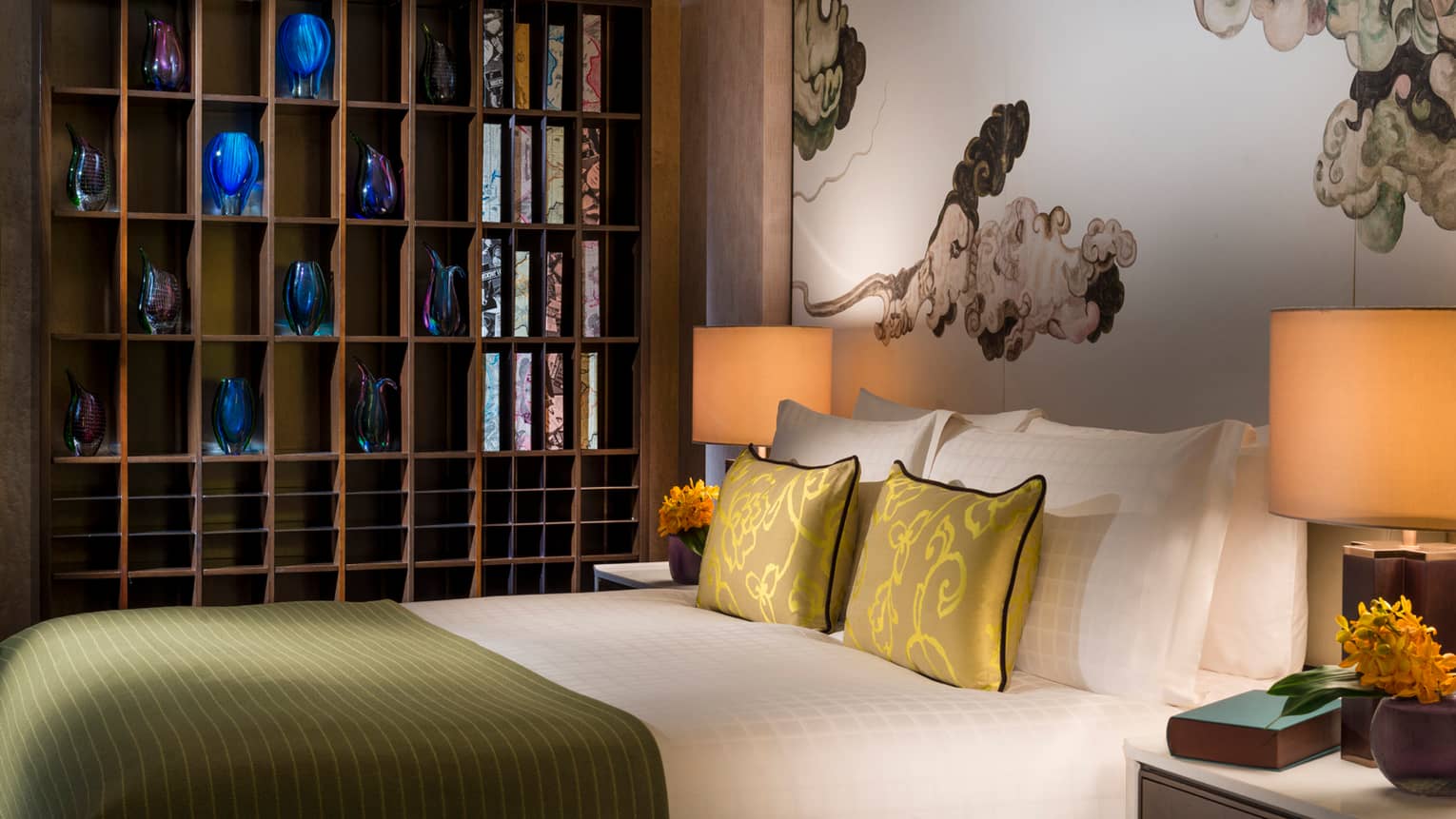 Hotel bed with gold silk pillows under wall shelf lined with blue, purple vases 