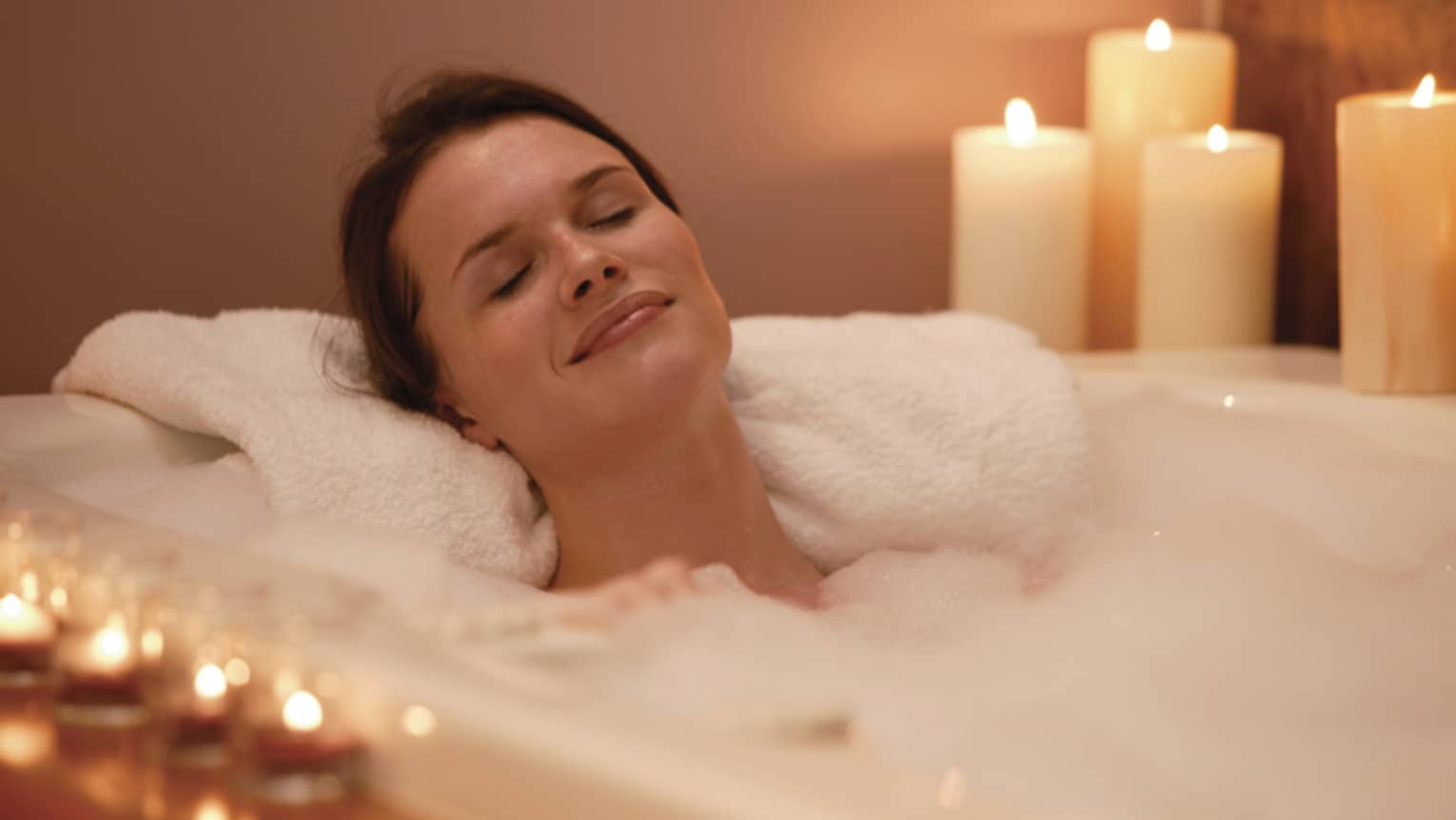 Woman with eyes closed relaxes in bubble bath with white candles on either side of tub