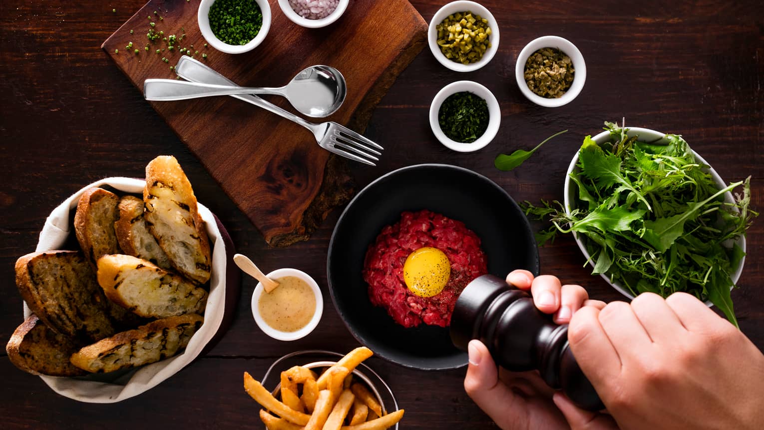 Hands grinding pepper over bowl of tartar, baskets with garlic toast, fries, lettuce, spices