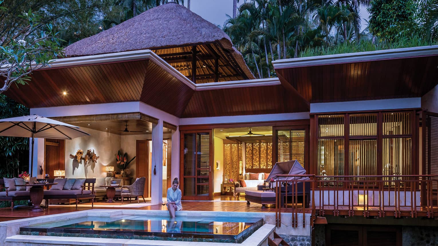 Woman dressed in white dips legs in pool on patio at night with large white-and-wood villa exterior in background