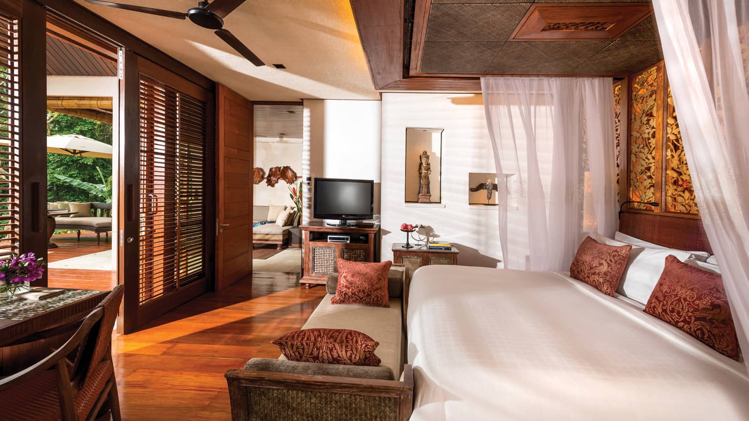 Villa bed with white linens, brown pillows, bench, small desk with fresh purple flowers, wood shutters open to patio