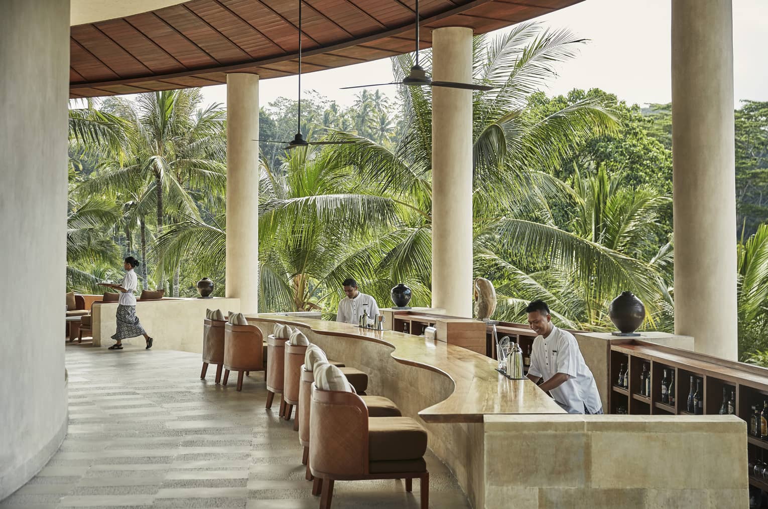 Smiling bartenders behind long wood-and-stone bar under white pillars of resort balcony, palm trees in background