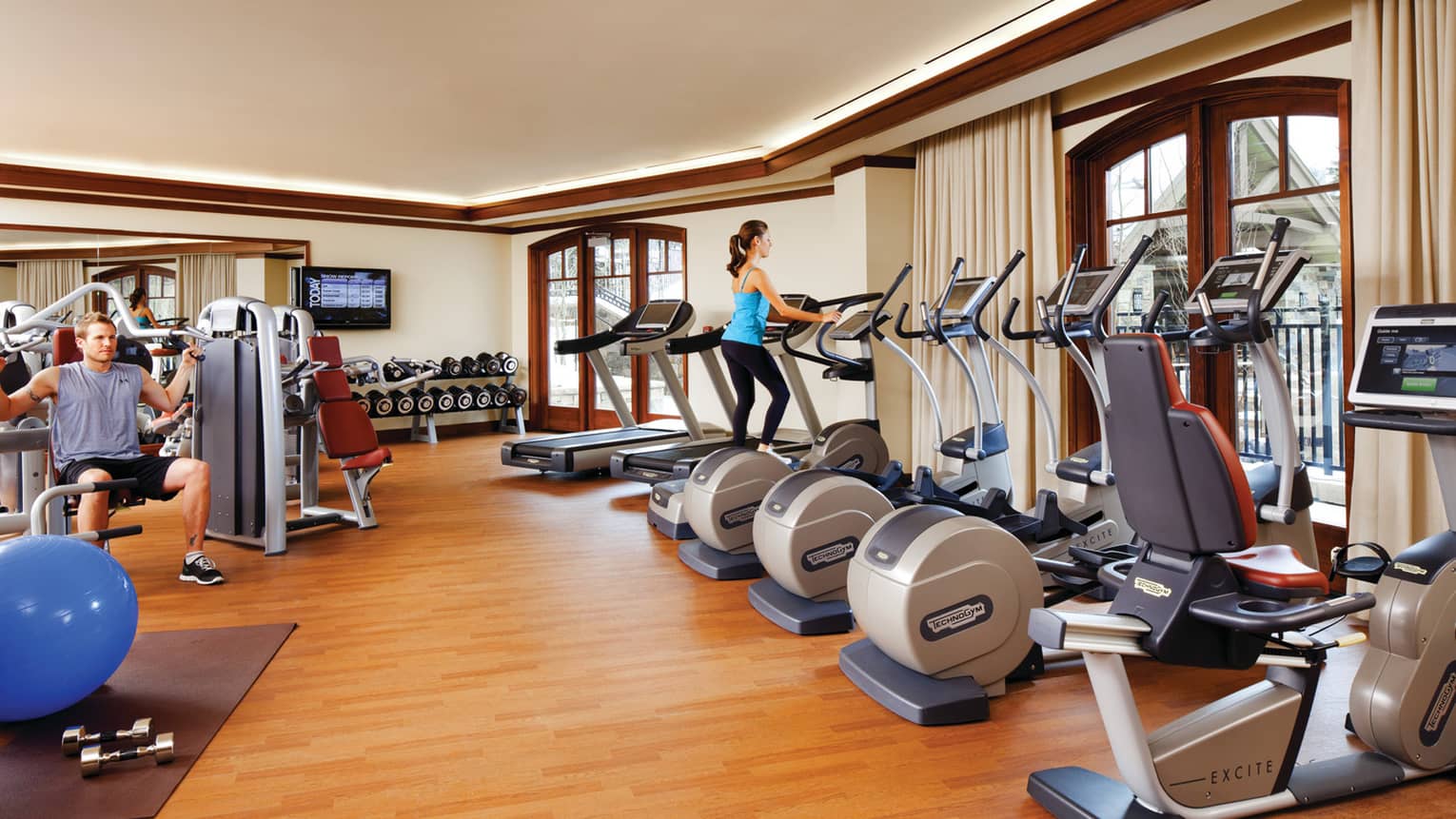 Woman on elliptical in row of cardio machines, man bench presses weights behind