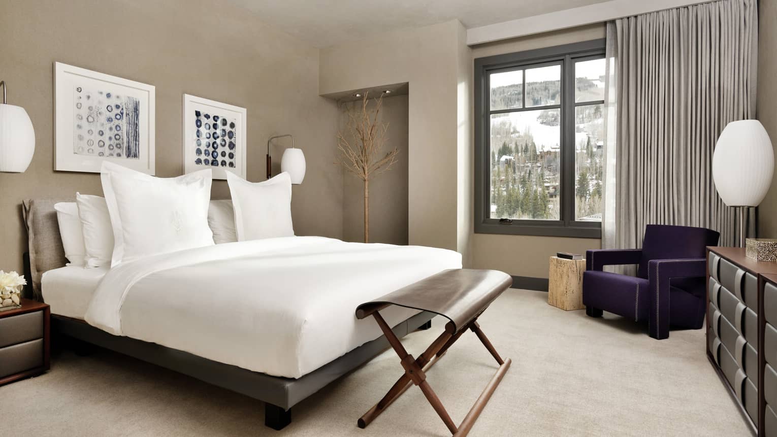 Bed with white linens, folding bench at foot, armchair by window with mountain views