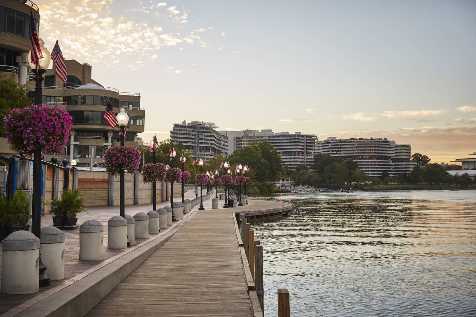 A waterfront with a wooden walkway next to light posts and flowers with building on the other side of a small road.