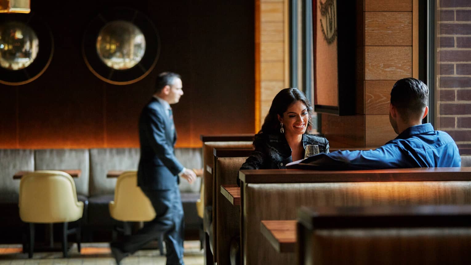 Smiling woman and man in booth in The Lounge at BOURBON STEAK dining room, man in suit walks in background