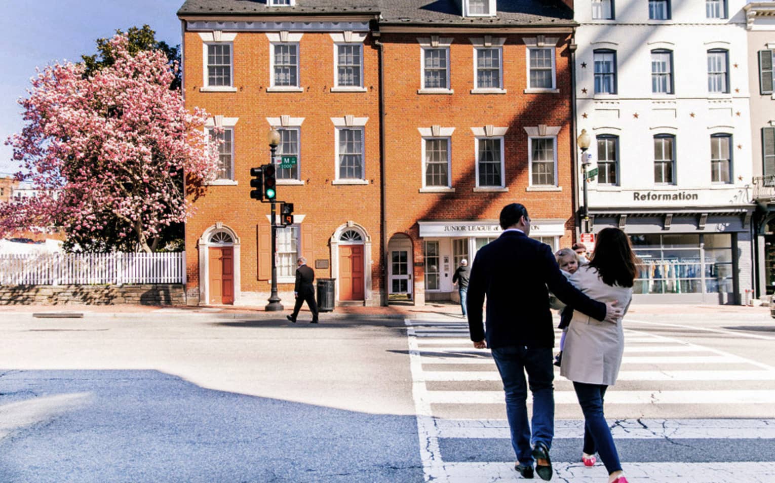 A man and woman walking along a cross walk towards an orange building with a cherry blossom tree next to it.