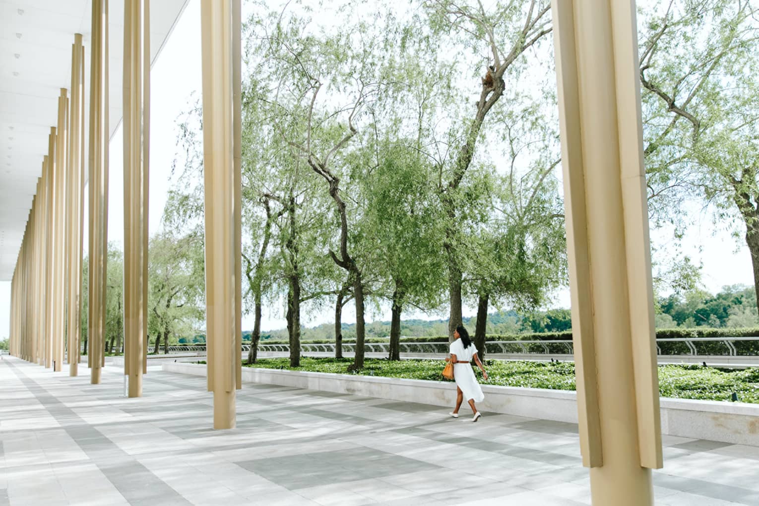 A woman inn a white dress holding an orange bag walking along beige columns outside near trees.