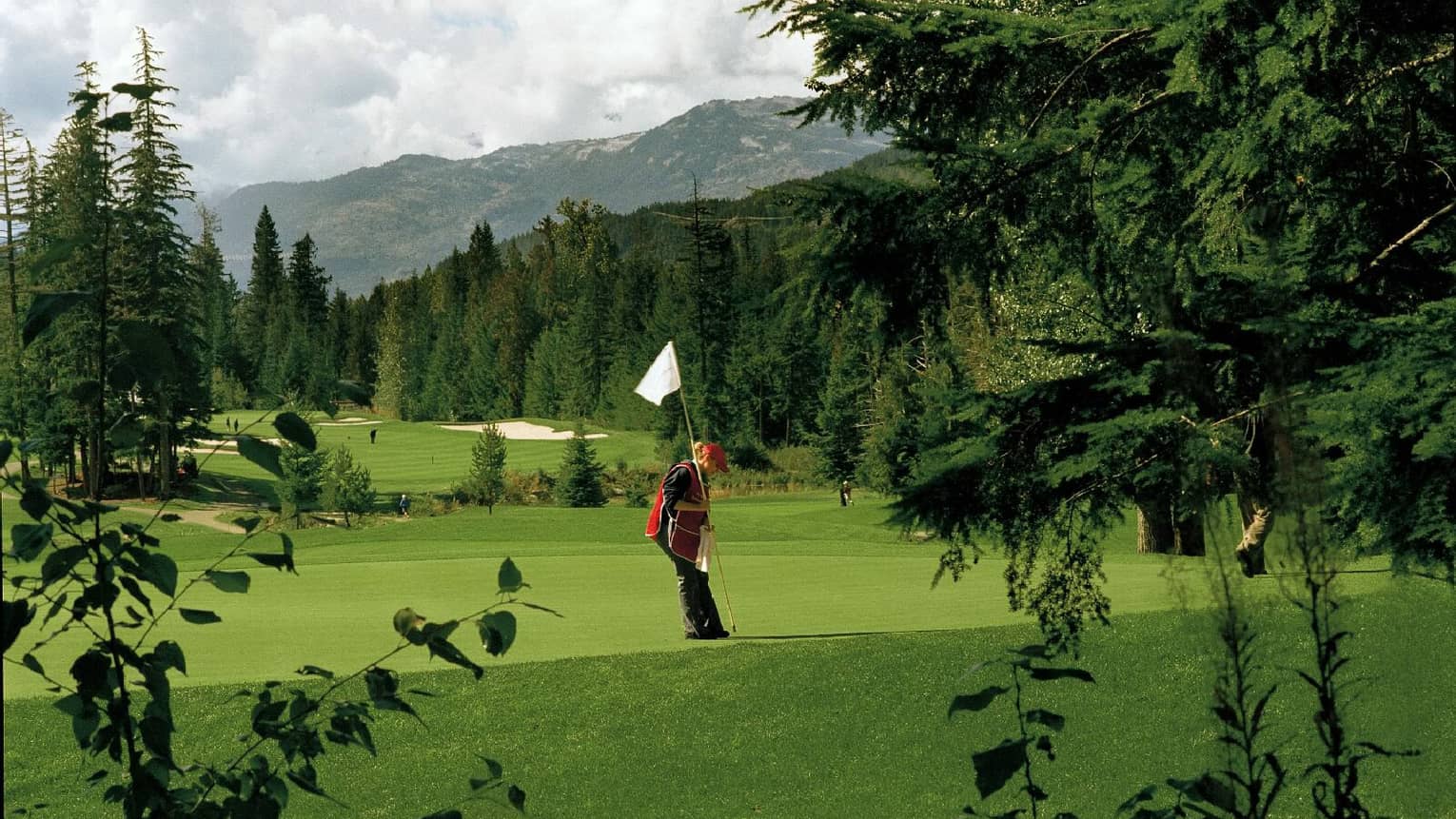 Woman wearing apron places pole with flag into hole on golf course green, mountains in background