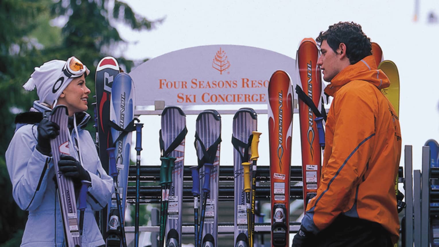 Man and woman in winter gear hold skis in front of Four Seasons ski rack