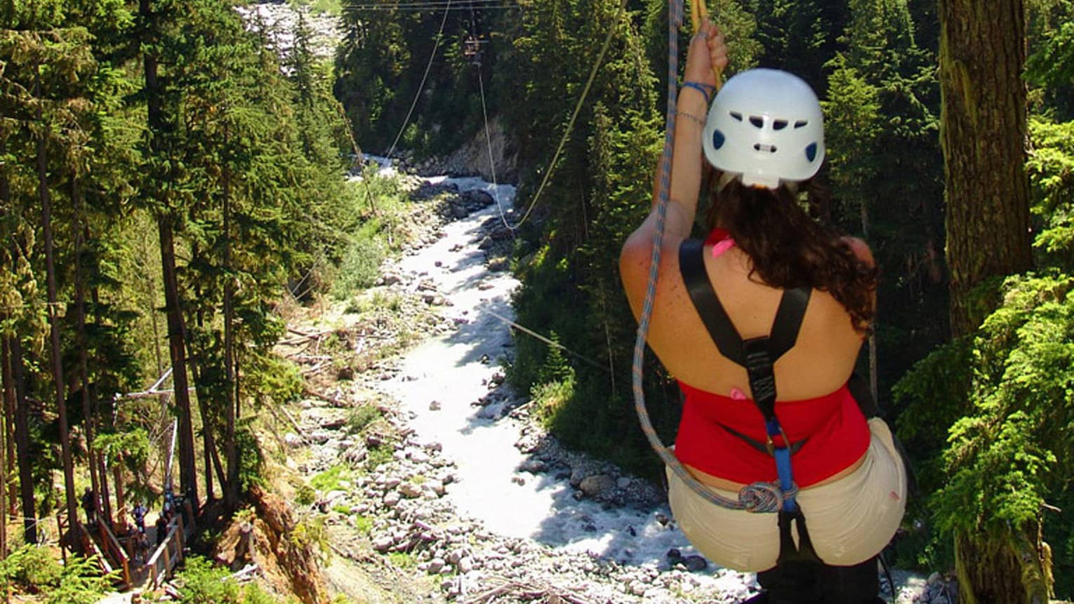 Back view of woman wearing helmet, hanging from zip line high above forest, creek