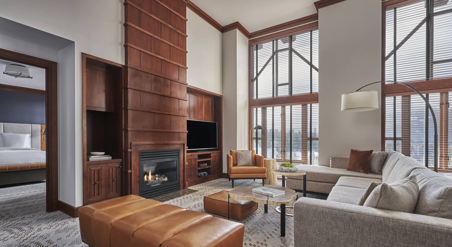 The Summit Suite at Four Seasons Whistler, featuring a luxurious and open interior with high ceilings and large, floor-to-ceiling windows that provide ample natural light and stunning views. The suite includes a cozy living area with a gray sectional sofa, a sleek glass coffee table, and a rich tan leather ottoman. A modern fireplace is set into a brick wall, adding warmth to the space. The bedroom area is visible in the background.,The Summit Suite at Four Seasons Whistler, featuring a luxurious and open interior with high ceilings and large, floor-to-ceiling windows that provide ample natural light and stunning views. The suite includes a cozy living area with a gray sectional sofa, a sleek glass coffee table, and a rich tan leather ottoman. A modern fireplace is set into a brick wall, adding warmth to the space. The bedroom area is visible in the background.