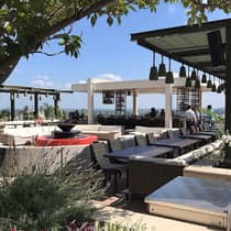 Outdoor rooftop lounge with modern seating, a red fire pit and bar area under a clear blue sky, surrounded by plants and shaded seating areas