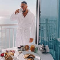 Man wearing white bath robe sips coffee in front of balcony breakfast table
