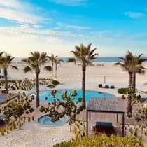 A tranquil beach scene with palm trees, a pool, a hot tub, and lounge chairs, set against a backdrop of white sand and a calm ocean under a clear blue sky.