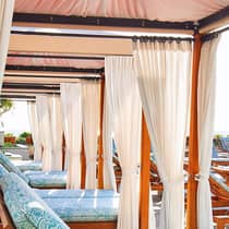 A row of patio lounge chairs with plush blue cushions under cabanas with white curtains, creating a luxurious and relaxing outdoor space.