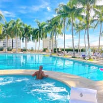Man in spa pool looks out over outdoor swimming pool, row of palm trees