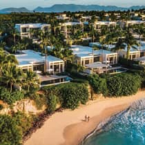 Aerial view of luxury resort beachfront villas surrounded by palm trees, with two people walking on the sandy beach below and mountains visible in the distance
