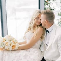 A bride and groom pose while sitting by a window overlooking a city skyline