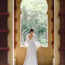 ,A woman in a white wedding dress stands in front of a large window overlooking green foliage