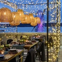 Tables with glassware and floral arrangements are set up under fairy lights on a patio overlooking the water and the Seattle Great Wheel.