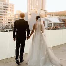 An opulent couple dressed in wedding attire hold hands on a rooftop overlooking the city skyline