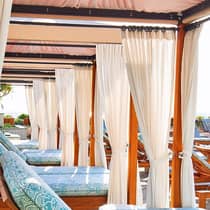A row of patio lounge chairs with plush blue cushions under cabanas with white curtains, creating a luxurious and relaxing outdoor space.