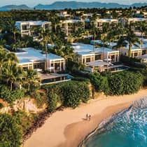 Aerial view of luxury resort beachfront villas surrounded by palm trees, with two people walking on the sandy beach below and mountains visible in the distance