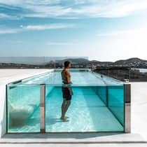 Man stands in raised glass swimming pool on white patio under blue sky
