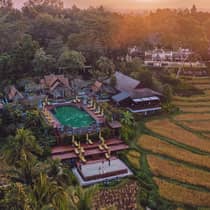 Aerial view of a resort with terraced pools, sun loungers and traditional architecture surrounded by rice fields at sunset.