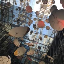 An artistic installation of floating abstract shapes suspended by wires, set against a backdrop of mirrored glass walls reflecting the sky and surrounding structures.