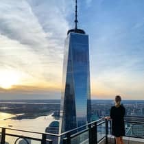 Person in a black dress standing on a rooftop terrace, overlooking One World Trade Center and the New York City skyline at sunset