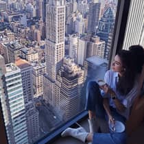 Woman with coffee cup sits in large window overlooking New York City rooftops