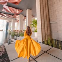 Person in a flowing yellow dress walking through a modern architectural space with tall columns, water features and a textured ceiling.