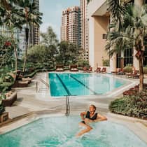 A person in black swimsuit relaxes in a jetted tub at an outdoor pool surrounded by lush greenery and tall residential buildings.