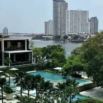 View of a modern building with a pool and palm trees in the foreground, overlooking a river with boats and tall city buildings across the water.